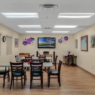 A well-lit communal room with tables, chairs, and a television mounted on the wall, decorated with artwork and purple floral designs.