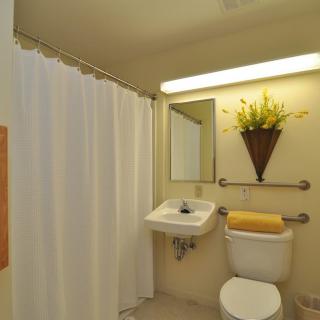 A neatly organized bathroom with a sink, toilet, and shower area. A mirror is mounted above the sink with decorative flowers and a towel placed on the toilet.