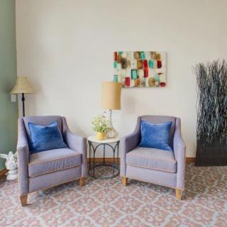 A cozy living room setup featuring two armchairs with blue throw pillows, a small round table with a lamp, artwork on the wall, and decorative plants.