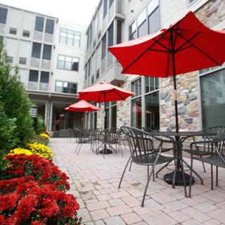 A cozy outdoor patio with red umbrellas, surrounded by flowers and adjacent to a modern building.