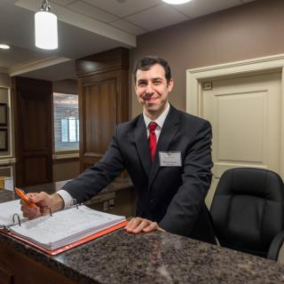 A receptionist standing at a hotel front desk, wearing formal business attire and smiling.