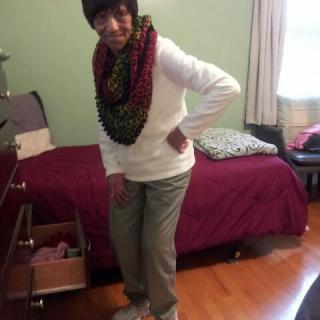 An elderly woman stands in a bedroom wearing a colorful scarf.