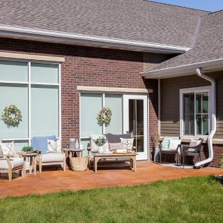 A cozy patio setup with outdoor furniture on a wooden deck attached to a brick house, featuring large windows and decorative wreaths.