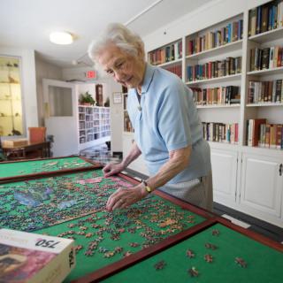 An elderly woman working on a puzzle in a room with bookshelves.
