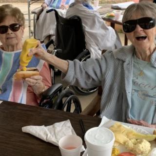 Two elderly women wearing sunglasses enjoy a meal together outdoors.