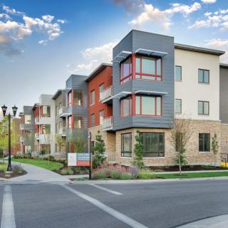 A modern apartment complex with well-maintained landscaping and contemporary architectural design, viewed from the street corner.