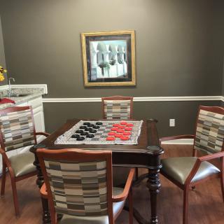 A cozy game room featuring a checkers set on a wooden table surrounded by chairs, with a painting adorning the wall.