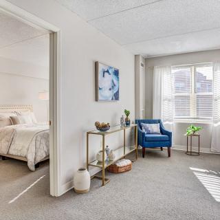 A cozy and stylish living space with an adjacent bedroom, featuring a blue chair, decorative elements, and soft natural light.