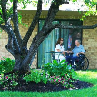 A caregiver assisting an elderly person in a wheelchair outside near a garden.