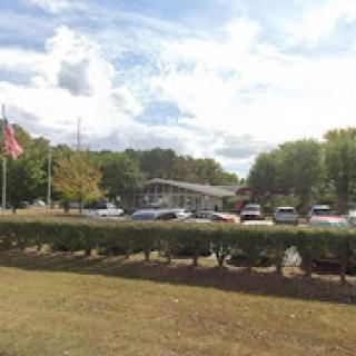 A view of a building with a parking lot, featuring a flag and surrounded by greenery under a cloudy sky.