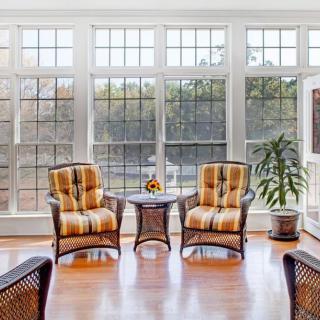 A sunlit room with two cushioned chairs and a small table, surrounded by large windows and a potted plant.