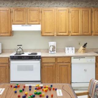 A cozy kitchen with wooden cabinets, a stove with a teapot, and various appliances on the counter. The dining table has colorful blocks scattered across it.