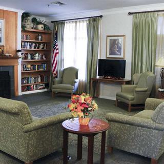 A cozy living room with a seating area, featuring armchairs around a small table with a flower arrangement. The room includes a fireplace, bookshelf, television, and is decorated with curtains.