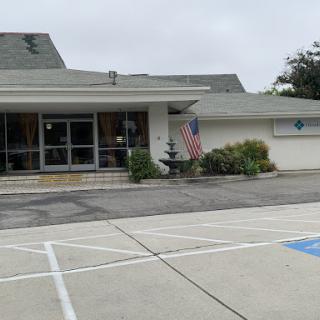A modest building featuring an American flag, surrounded by a parking lot and greenery under a cloudy sky.