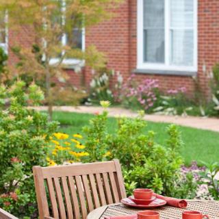 A garden scene with a wooden patio table set for dining, surrounded by vibrant flowers and greenery in front of a brick house.