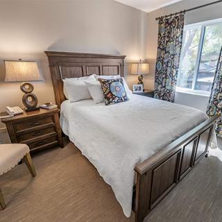 A cozy bedroom with a wooden bed frame, white bedding, and accompanying nightstands with lamps. The room is lit by natural light from a window adorned with floral curtains.