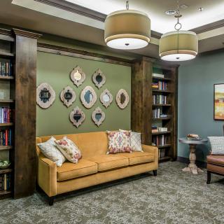 A cozy living room with a mustard-colored sofa, surrounded by bookshelves and decorative artwork on the walls.