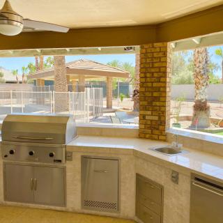 A spacious outdoor kitchen with a barbecue grill and brick pillars overlooking a sunny backyard with palm trees.