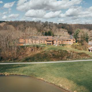 A serene view of a multi-story building nestled among trees, with a pond in the foreground and a cloudy sky above.