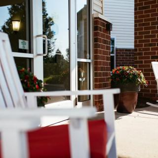 A sunny patio area with white chairs and potted plants outside a brick building.