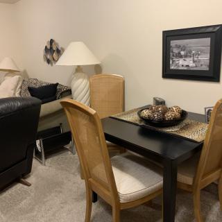 A cozy living room with a small dining area, featuring wooden chairs, a table, lamps, and wall artwork.