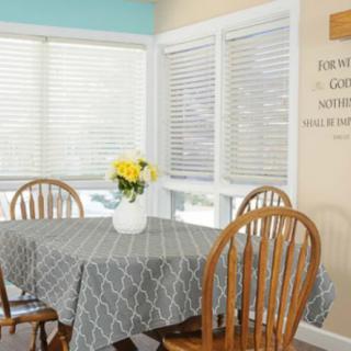 A cozy dining area with a round table covered by a gray patterned tablecloth, surrounded by wooden chairs. A vase with yellow flowers sits at the center, and window blinds are drawn behind it.