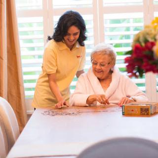 A caregiver and an elderly woman engage in a warm activity at a table, fostering companionship and connection.
