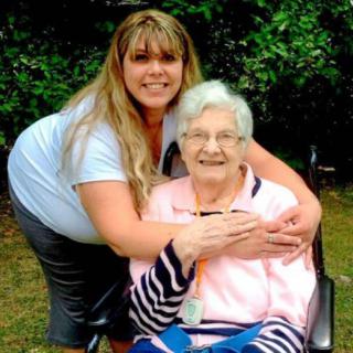 A young woman embraces an elderly woman in a wheelchair, both smiling while enjoying time outdoors.