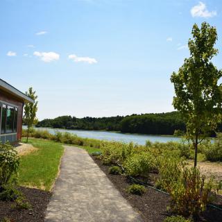 A scenic view of a pathway leading alongside a lake with trees and a building nearby.