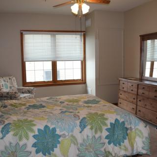 A cozy bedroom with a floral bedspread, wooden dresser, and large windows with blinds.