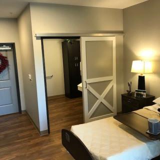 A cozy bedroom with wooden flooring, featuring a bed, nightstand with a lamp, and a sliding door.