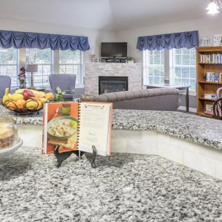 A cozy kitchen with a cookbook on the countertop, overlooking a living room with a fireplace and bookshelf.