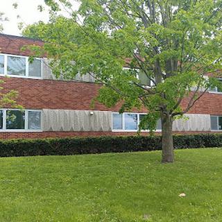 A brick building with several windows, surrounded by a grassy area and trees.