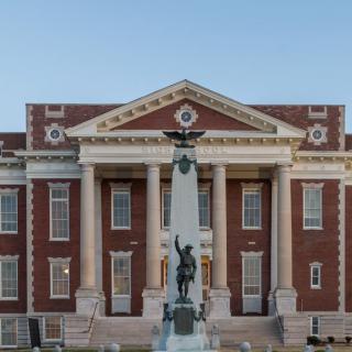 A historical red brick building with classical columns and a monument in front.