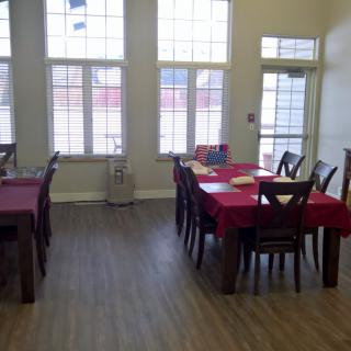 A bright dining room with tables covered in red tablecloths, surrounded by wooden chairs and illuminated by large windows.