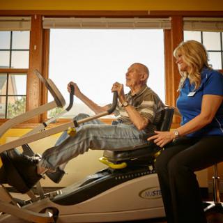 A fitness trainer assists an elderly man as he uses a specialized exercise machine in a rehabilitation setting.