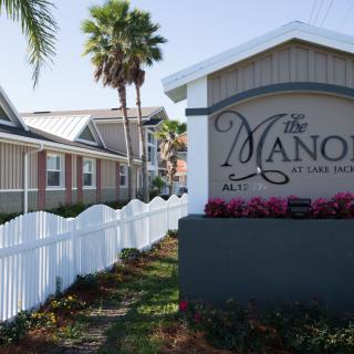 A row of houses with a white picket fence and palm trees, featuring a sign for 'The Manor'.