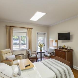A cozy bedroom with a bed, chairs, and a television, featuring modern decor and natural light from the windows.