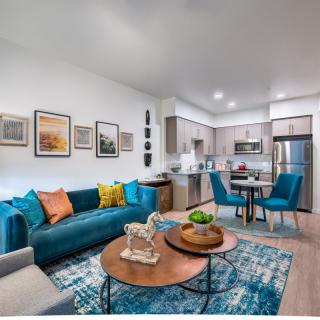 A modern open-concept living area with a blue sofa, colorful pillows, and a kitchen in the background.