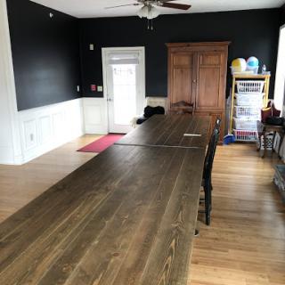 A spacious interior room featuring a long wooden table, black walls, and a warm wooden floor.