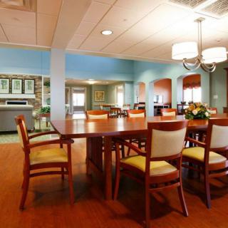 A well-lit dining room with a large wooden table and chairs, featuring a kitchen area in the background.