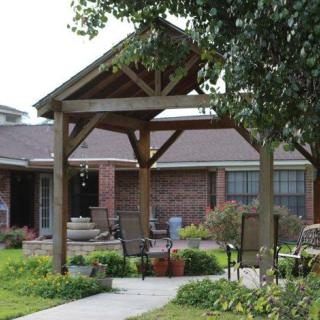 A cozy garden patio area with a wooden pergola and outdoor seating in front of a brick house.