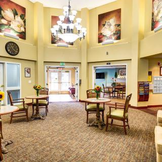 A spacious and well-lit lobby area featuring tables, chairs, a chandelier, and decorative paintings.