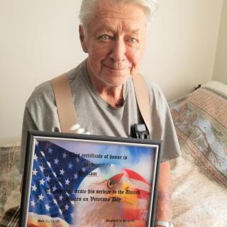 An elderly man proudly holds a framed award featuring an American flag.
