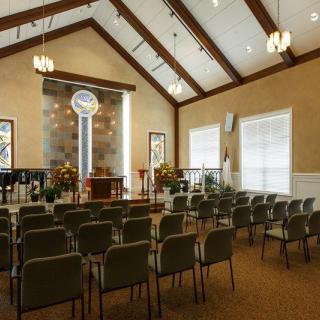 An interior view of a chapel with rows of seating facing an altar, featuring large windows and decorative lighting.