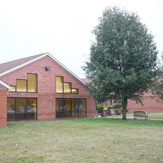 A brick school building with large windows, a tree, and a bench in the foreground.