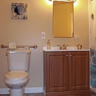 A bathroom featuring a toilet, sink with a wooden cabinet, and a mirror above. There's artwork on the wall and a towel rack beside the toilet.