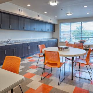 A modern kitchen and dining area with orange chairs and tables, featuring ample natural light from large windows.