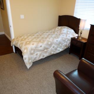 A cozy bedroom featuring a single bed with a patterned comforter, a nightstand with a lamp, and an upholstered chair.