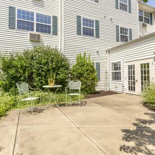 A sunny patio area with a small table and chairs, surrounded by lush greenery next to a residential building.